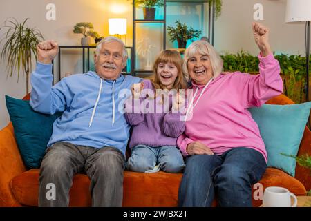 Glückliche kaukasische Großeltern und Enkelin schauen im Fernsehen schreiend, feiern Erfolg, Sieg, Zielerreichung gute Nachrichten, Sieg sitzen auf Sofa im Wohnzimmer. Familie ballt zu Hause Fäuste. Stockfoto