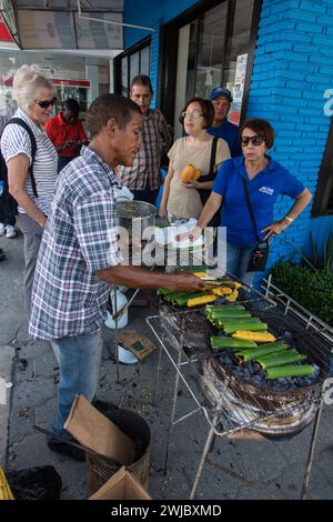 Ein Mann kocht dominikanische Tamales auf einem offenen Grill auf der Straße auf der Bani Mango Expo in Bani, Dominikanische Republik. Ein attraktiver blonder Seniorentouris Stockfoto