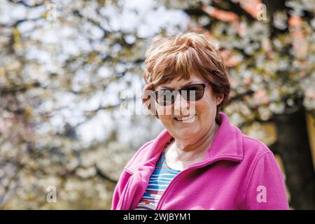 Porträt einer glücklich lächelnden Seniorin unter blühendem Kirschbaum im Garten im Frühling. Ältere Person genießt den sonnigen Frühlingstag Stockfoto