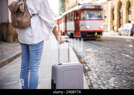 Frau, die auf der Straße auf die Straßenbahn wartet, mit Koffer und Rucksack. Fahrt mit öffentlichen Verkehrsmitteln in der Stadt Stockfoto
