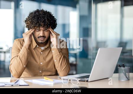 Müder und kranker junger indianischer Mann, der im Büro am Schreibtisch sitzt und grimmig, unter schweren Kopfschmerzen und Druck leidet und seinen Kopf mit den Händen massiert. Stockfoto