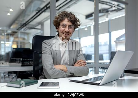 Ein professioneller junger Geschäftsmann mit lockigem Haar, der selbstbewusst an seinem Laptop in einer modernen Büroumgebung arbeitet. Stockfoto
