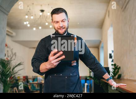 Lächelnder Inhaber eines kleinen Unternehmens in schwarzer Chefuniform mit Schürze und digitalem Tablet in seinem gemütlichen Restaurantsaal. Erfolgreiche Menschen, hart W Stockfoto