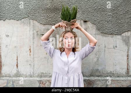 Lustige blonde Frau, die einen Topf mit Pflanzen in Bastelpapier auf dem Kopf hält Stockfoto