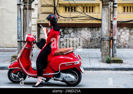 Die stilvolle junge vietnamesische Frau hält auf der Straße an, während sie mit einem Vespa-Roller fährt, um ihr Handy zu überprüfen. Hanoi, Vietnam. Stockfoto