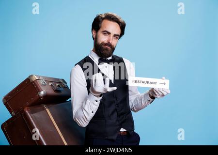 Hotelangestellter hält ein Restaurantschild, um Touristen mit Wegbeschreibungen zu helfen, und posiert mit dem Bereichszeiger auf der Kamera. Junger, klassischer Pagenjunge zeigt ein Symbol für den Speisesaal. Stockfoto