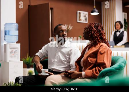 Leute mit Zimmerreservierung in der Lobby, sitzen im Loungebereich und unterhalten sich. afroamerikanische Hotelgäste, die an der Rezeption auf die Anmeldung warten, luxuriöse, moderne Unterkünfte. Stockfoto