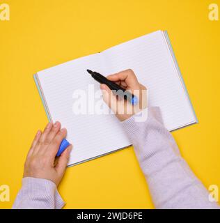 Ein offenes Notizbuch mit leeren weißen Blättern und einer Frauenhand, die einen Filzstift auf gelbem Hintergrund hält, Draufsicht Stockfoto