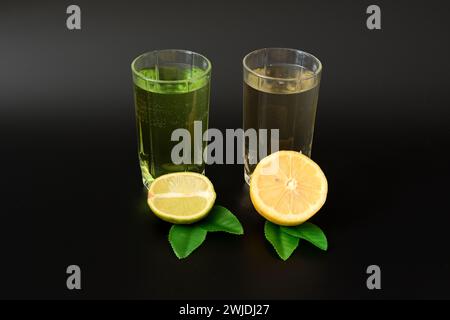 Zwei Gläser mit verschiedenen Zitrussäften auf schwarzem Hintergrund stehen in einer Reihe mit Hälften reifer Limetten und Zitronen mit Blättern. Nahaufnahme. Stockfoto