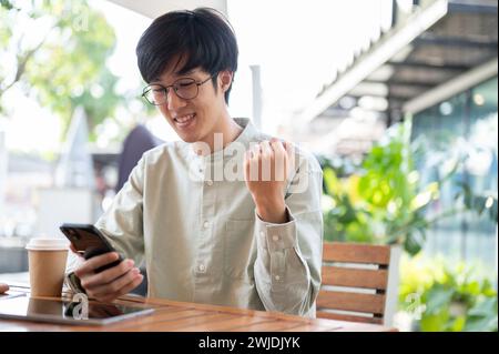 Ein glücklicher asiatischer Mann erhält gute Nachrichten über seine Nachricht, indem er sein Smartphone mit fröhlichem Gesicht ansieht, während er in einem Café sitzt. Stockfoto