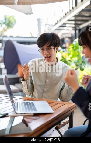 Zwei begeisterte junge asiatische Kollegen feiern gemeinsam ihren Projekterfolg, schauen auf einen Laptop-Bildschirm mit überraschten Gesichtern und heben ihre f Stockfoto