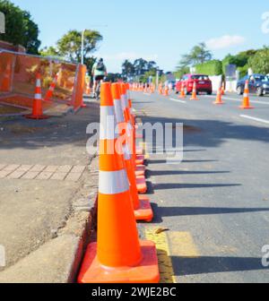 Orangefarbene Verkehrskegel, die die Straße entlang Reihen. Nicht wiedererkennbare Radfahrer und Autos auf der Straße. Bauarbeiten in Auckland. Stockfoto