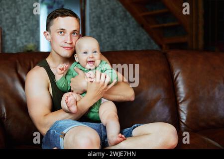 Junger Europäischer Vater hält seine kleine Tochter in den Armen, die lächelt und ihre Zunge zeigt, Familie sitzt zu Hause auf dem Ledersofa im Wohnzimmer. Stockfoto