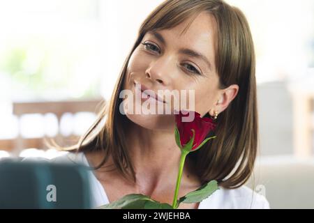 Eine junge kaukasische Frau lächelt sanft, während sie eine rote Rose nahe an ihr Gesicht hält Stockfoto