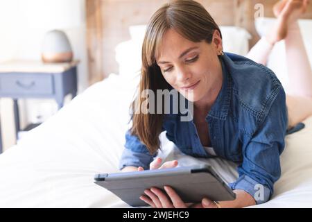 Eine weiße Frau mittleren Alters liegt auf einem Bett, während sie sich mit einer Tablette beschäftigt Stockfoto