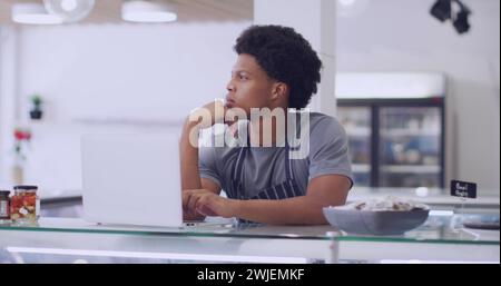 Ein junger afroamerikanischer Mann denkt über seinen Laptop in einer Küche nach Stockfoto
