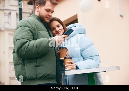 Wunderschönes, schönes Paar, das Kaffee auf der Terrasse trinkt und ein Mann, der das Telefon durchblättert, während er nach Lieferservice oder Geschäften im Internet sucht. Stockfoto