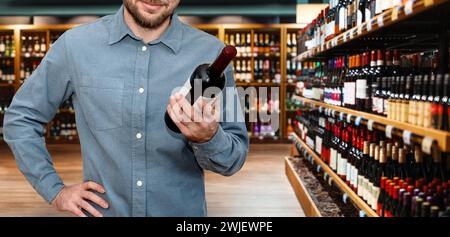 Man Kunde wählt den Wein im Spirituosengeschäft. Männliche Person, die eine Rotweinflasche in der Hand hält, auf dem Hintergrund des Weingeschäfts. Stockfoto