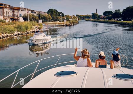 Aigues-Mortes (Südfrankreich): Tourismus auf dem „Kanal der Rhone a Sete“ (Kanal der Rhone-Sete). Drei Frauen, die von hinten auf einem Mietkahn gesehen werden, winken als Stockfoto