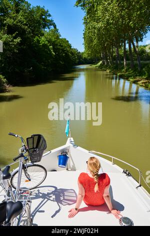 Binnenschifffahrt auf dem Canal du Midi. Frau, die sich entspannt und die Sonne auf dem Vordeck genießt, neben ihrem Fahrrad. Der Canal du Midi ist als UNESCO Wor eingetragen Stockfoto