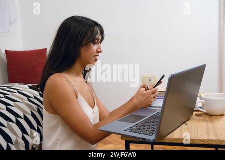 Seitenansicht einer jungen brünetten Latina-Frau zu Hause, die auf dem Boden sitzt, mit einem Laptop arbeitet und das Handy überprüft, um Nachrichten von ihren Kunden zu lesen. Kopieren Stockfoto