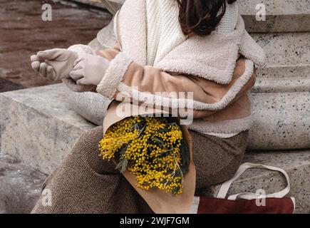 Modische Details aus beigefarbener warmer Jacke, braunem Rock und einem Blumenstrauß frischer Mimosa. Stylischer Winter-Look Stockfoto