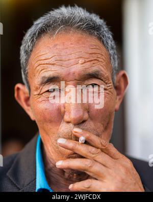 Ein Porträt eines älteren Ladakhi-Mannes, der eine Zigarette raucht und in die Kamera schaut. Stockfoto