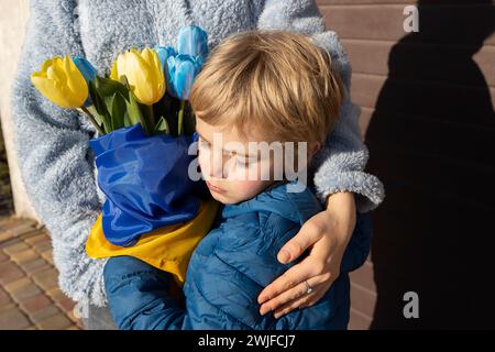 Trauriger kleiner Junge umarmt eine Frau, die einen Strauß aus gelben und blauen Tulpen in der Hand hält, die in eine ukrainische Flagge gewickelt sind. Familie, Zweisamkeit, Unterstützung. Stockfoto