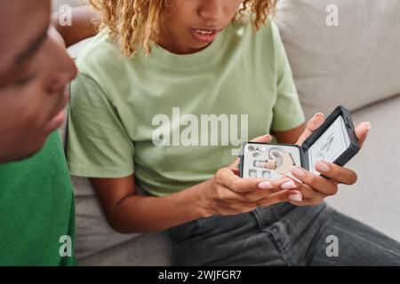 Abgeschnittene afroamerikanische Frau, die Hörgerät in einer Kiste neben dem Freund hält, während sie auf der Couch sitzt Stockfoto