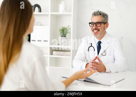Ein älterer männlicher Arzt mit grauen Haaren und Brille wird eine Patientin beruhigend anlächeln sehen, was eine warme und einladende Atmosphäre in einem modernen, gut beleuchteten Sprechzimmer schafft. Stockfoto