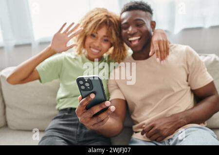 Konzentrieren Sie sich auf Smartphone, fröhliches afroamerikanisches Paar, das im Wohnzimmer chattiert und winkt Stockfoto
