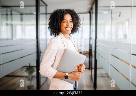 Eine selbstbewusste brasilianische Geschäftsfrau mit lockigen Haaren hält ihren Laptop und lächelt in die Kamera. Sie steht in einem modernen Bürosaal. Das Bild vermittelt ein Gefühl des beruflichen Erfolgs und der Zugänglichkeit Stockfoto