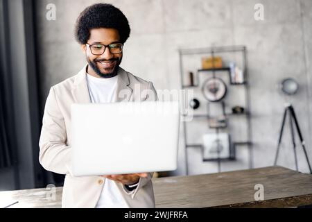 Ein junger, stilvoller Geschäftsmann in einem beigefarbenen Blazer hält einen Laptop und drückt sein Gesicht Konzentration und Professionalität aus. Die moderne Büroeinrichtung im industriellen Stil trägt zur modernen Atmosphäre bei Stockfoto