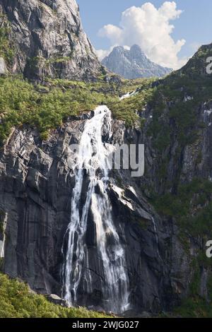 Der Valefossen-Wasserfall fließt kräftig die Klippen der Lofoten-Inseln in Norwegen hinunter, inmitten üppiger Vegetation und unter einem dynamischen Sommerhimmel. Stockfoto