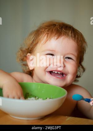 Lustige Baby Essen selbst mit einem Löffel in der Küche. Lächelndes Baby beim Essen. Stockfoto