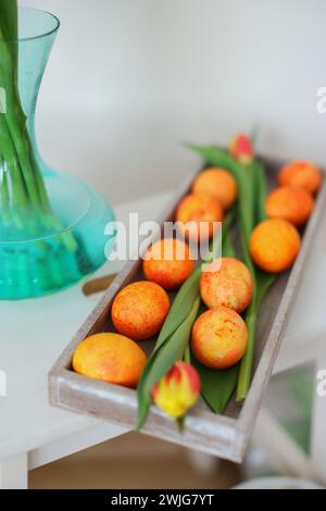 Frohe Ostern, stilvolle Ostereier auf Holzteller, Tulpen und Leinenserviette auf rustikalem weißen Tisch Stockfoto