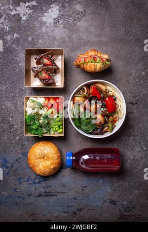 Köstliches Mittagsmenü, Pasta, Salat, Sandwich, Bagel, Saft auf Steintisch. Pasta mit Gemüse, frischer Salat, Schinken Käse Sandwich, weicher Bagel, Saft. Stockfoto