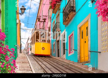 Gelbe elektrische Straßenbahn auf einer Straße mit bunten Häusern und Blumen auf den Balkonen - Bica Aufzug, der den Hügel von Chiado in Lissabon, Portugal, hinunter fährt. Stockfoto