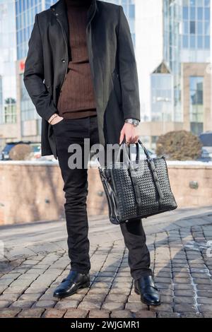 Geschäftsmann in schwarzem Mantel, Rollkragenpullover, Ledertasche in der Hand, in der Nähe des Büros. Tragen von Jeansschuhen. Niedriger Winkel, unscharfer Hintergrund. Stockfoto