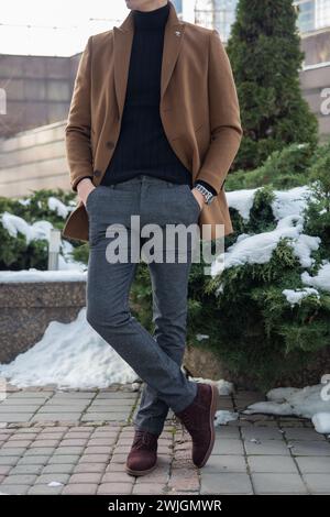 Mann in braunem Mantel, schwarzem Rollkragen, grauen Hosen und braunen Schuhen, der vor einem Gebäude mit Händen in Taschen steht. Stockfoto