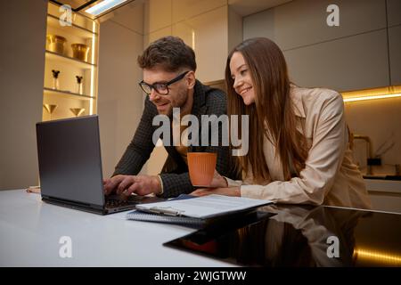 Junge Familie, die Laptop in der Küche benutzt, Ehemann hilft der liebenden Frau mit Projekt Stockfoto