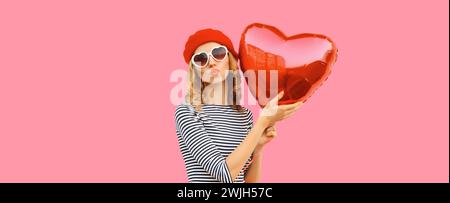 Süßes Porträt der schönen glücklichen jungen Frau mit rotem Herzballon bläst Kuss im französischen Baskenmütze auf rosa Studiohintergrund Stockfoto
