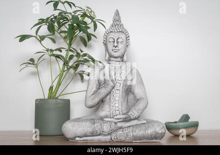 Dekorative Buddha-Statuette in meditativer Position auf einem Holztisch, neben einem Topf mit Pflanzen und einer kleinen Schüssel mit grünem Stein. Weißes Ba Stockfoto