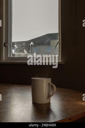 Ein einsamer weißer Kaffeetasse sitzt auf einem Holztisch am Fenster des Apartments im Morgenlicht. Morgenkaffee, das Konzept der Einsamkeit und Stockfoto