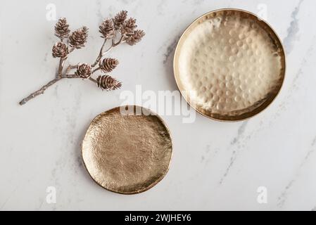 Zwei goldene Teller und ein dekorativer goldener Zweig mit Tannenzapfen auf weißem Marmorhintergrund, Blick von oben Stockfoto