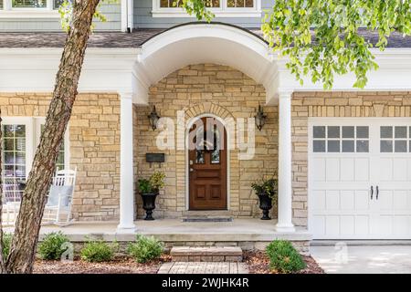 Ein Vordertür-Detail mit einer abgerundeten Fassade und Veranda, einem Stein-und blauen Abstellgleis und einem weißen Garagentor mit Fenstern. Stockfoto