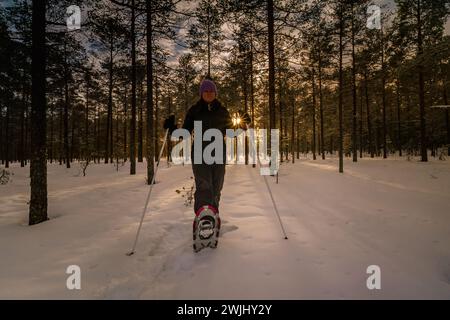 Frauen mittleren Alters in Schneeschuhen posieren im Kiefernwald. Die Abendsonne scheint durch Bäume im Hintergrund Stockfoto