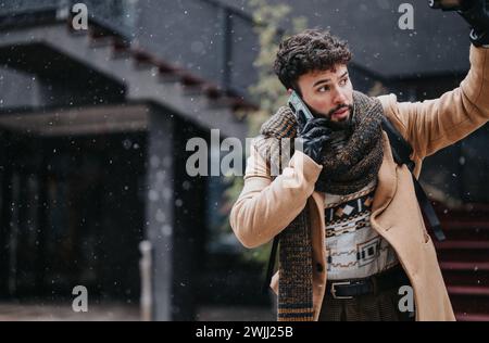 Ein fokussierter junger Geschäftsmann in stilvoller Winterkleidung führt ein ernsthaftes Telefongespräch, während Schneeflocken sanft um ihn fallen. Stockfoto