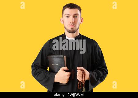 Junger Priester mit der Heiligen Bibel und betenden Perlen auf gelbem Hintergrund Stockfoto