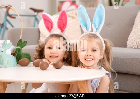 Niedliche kleine Mädchen in Hasenohren mit Schokoladen-Ostereiern auf dem Tisch zu Hause Stockfoto